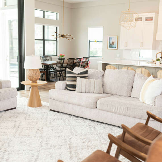 light livingroom with white carpet and wood and leather chaires, white lamp on top of wood small table with white kitchen in the back with marble finishes